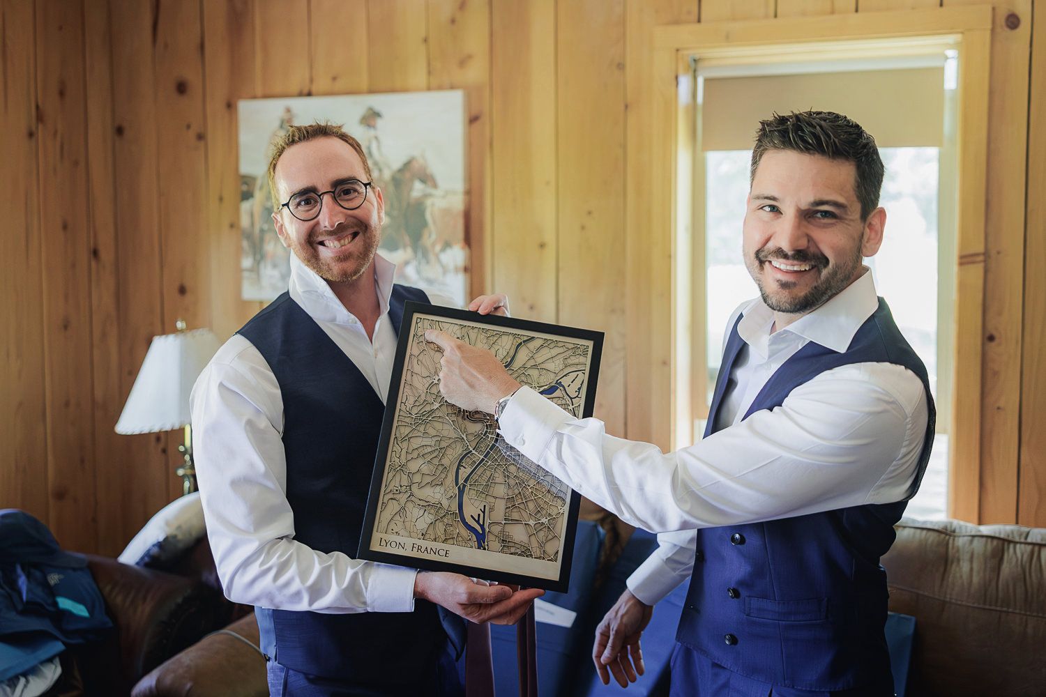 Two men in waistcoats smiling while holding and displaying a framed piece of artwork in a wood-paneled room.
