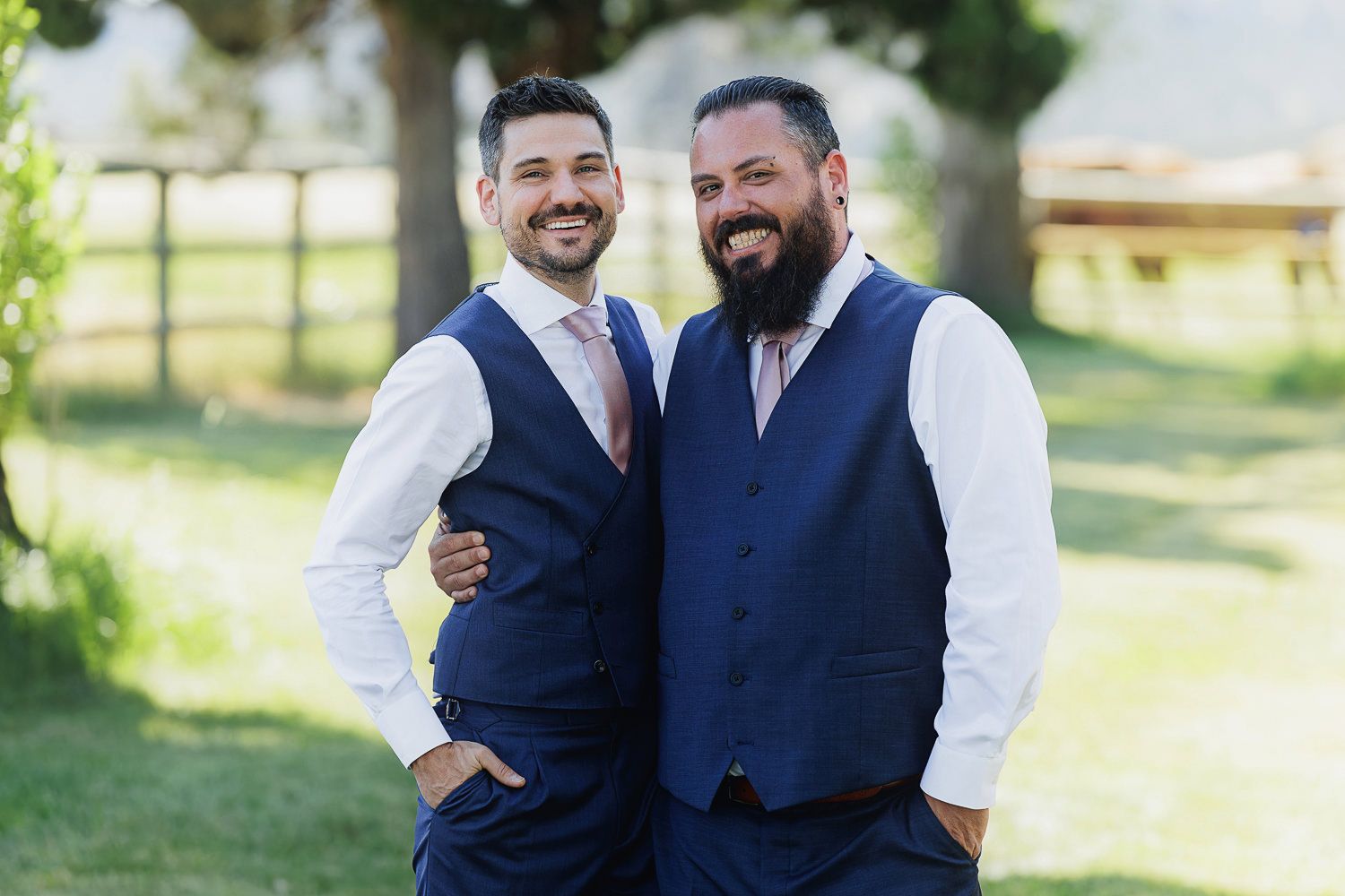 Two people in matching navy vests and white dress shirts embrace outdoors on a sunny day.