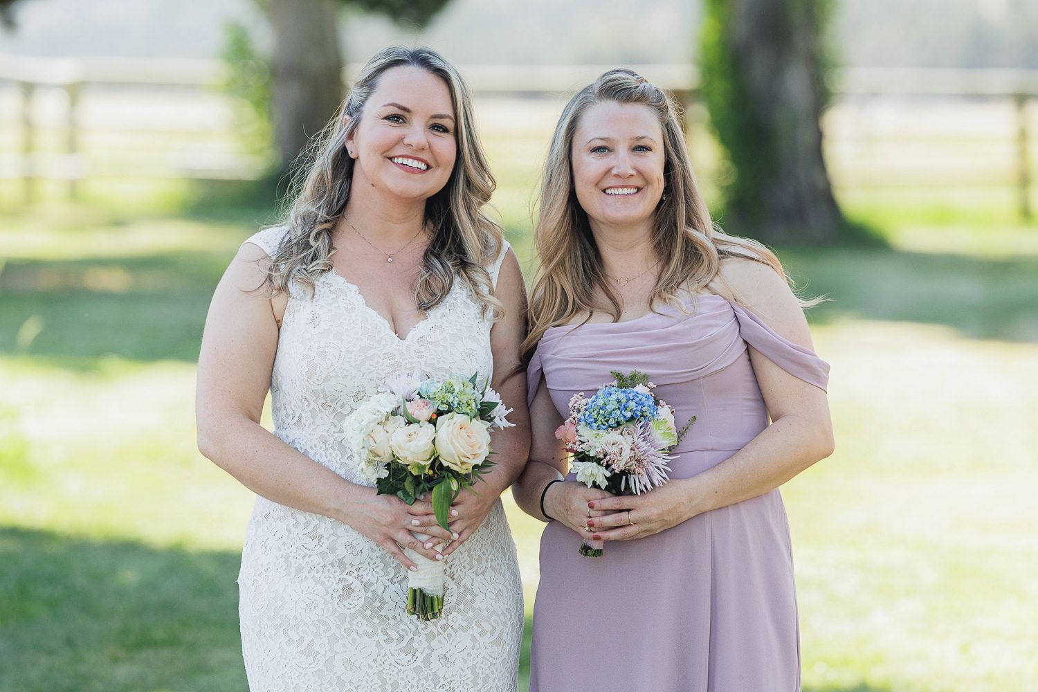 Two women in wedding attire holding bouquets smile together outdoors on a sunny day.