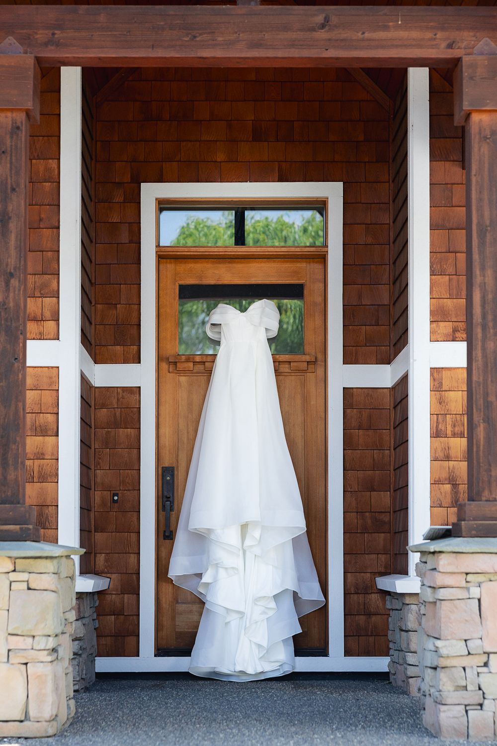 A flowing white wedding dress hangs elegantly on a wooden door with stone pillars and cedar shingle siding.