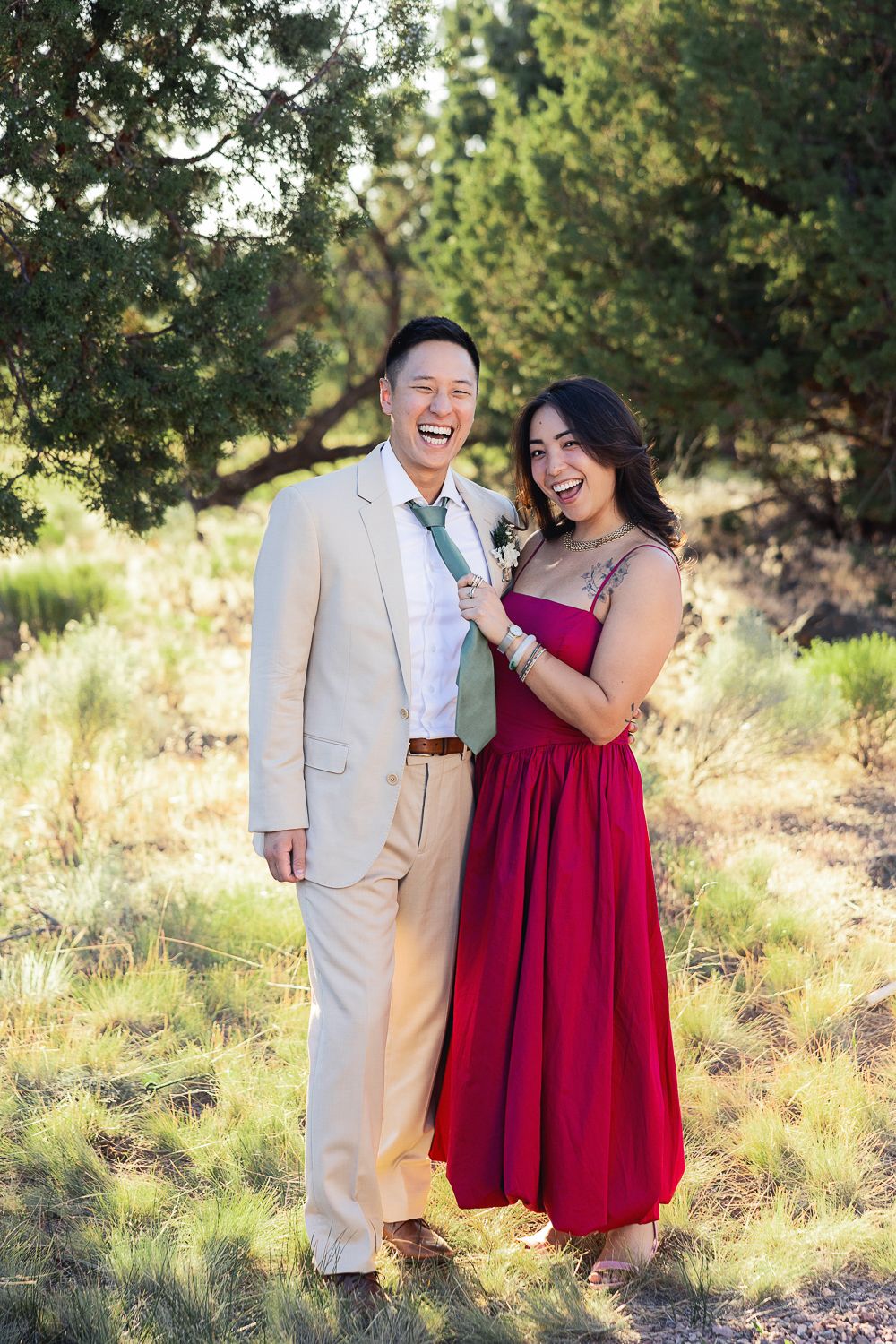 Couple in red dress and beige suit laughing together in a natural outdoor setting.