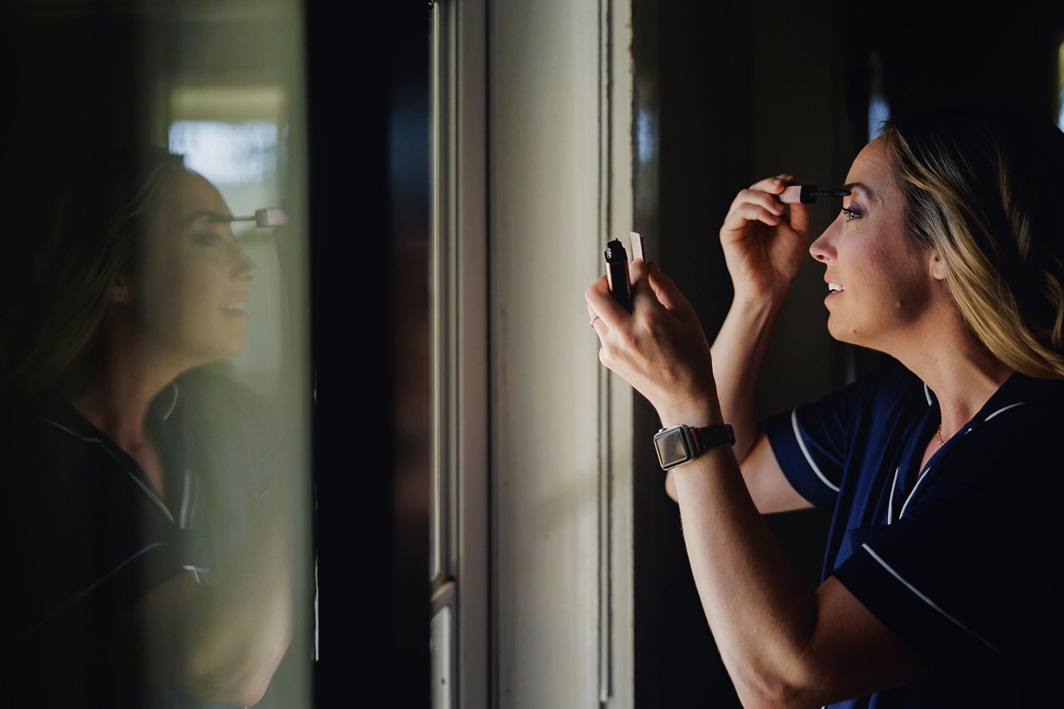 Person applying mascara while looking in window reflection during golden hour sunlight.