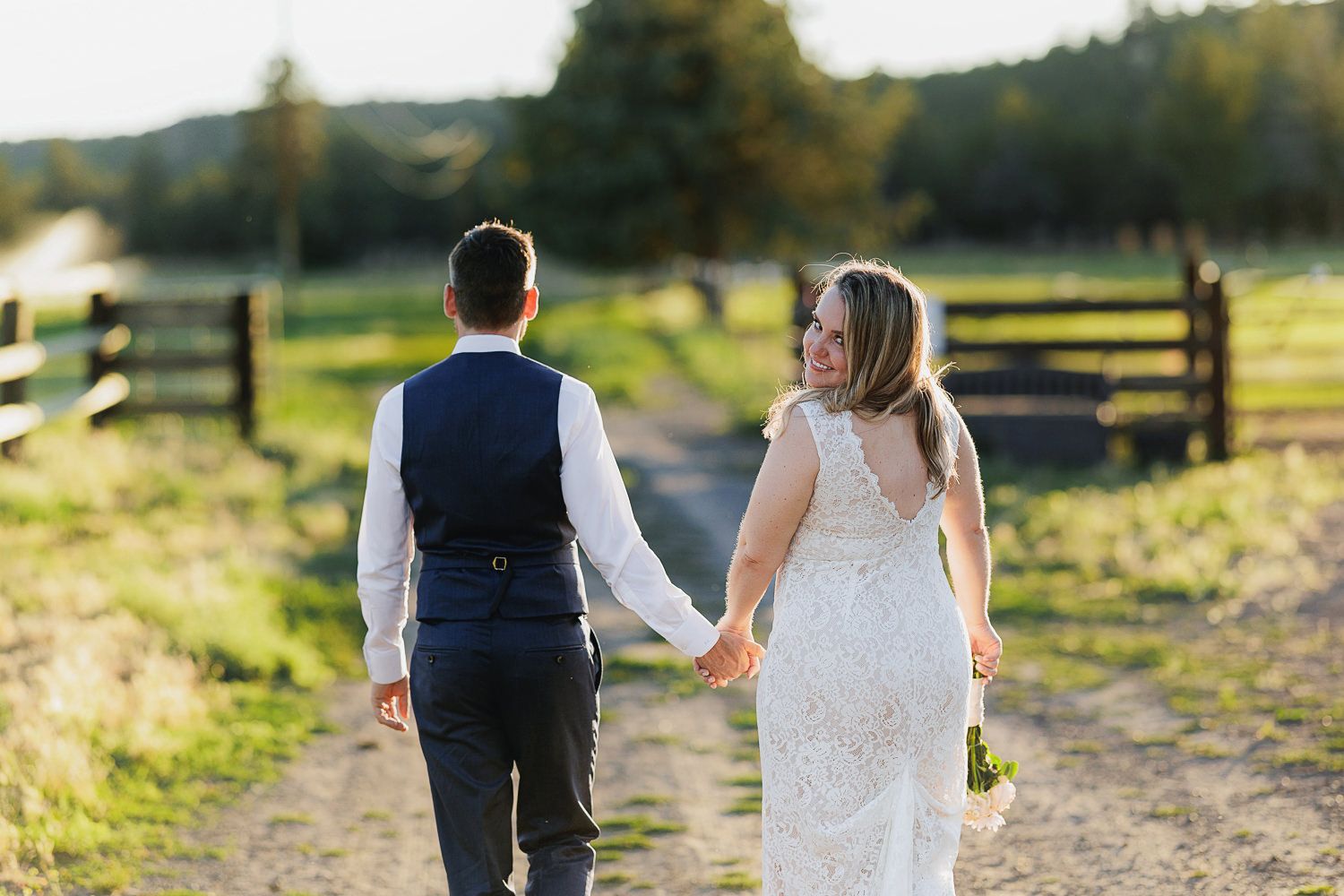 Newlywed couple holding hands walking down a sunlit country path with wooden fences and green fields.
