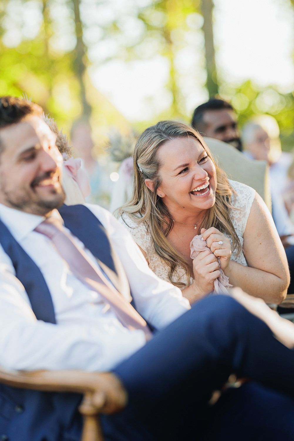 Two wedding guests share a lighthearted moment together while seated outdoors with natural lighting and bokeh effect.