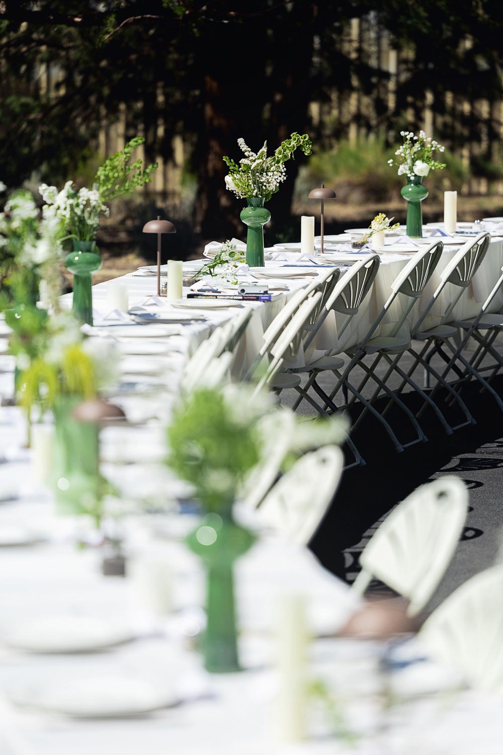 An elegant outdoor table setting with white linens, place settings and green floral centerpieces in tall vases.