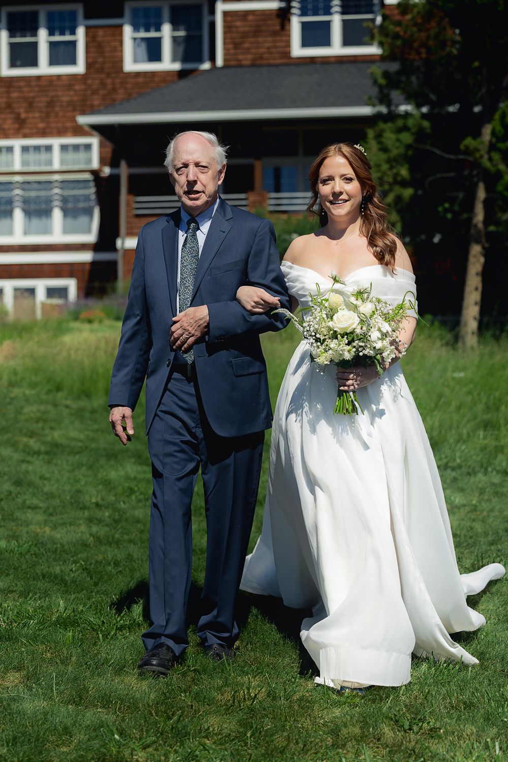 A bride in a white off-shoulder gown carrying a bouquet walks arm in arm with someone in a navy suit on a grassy lawn.