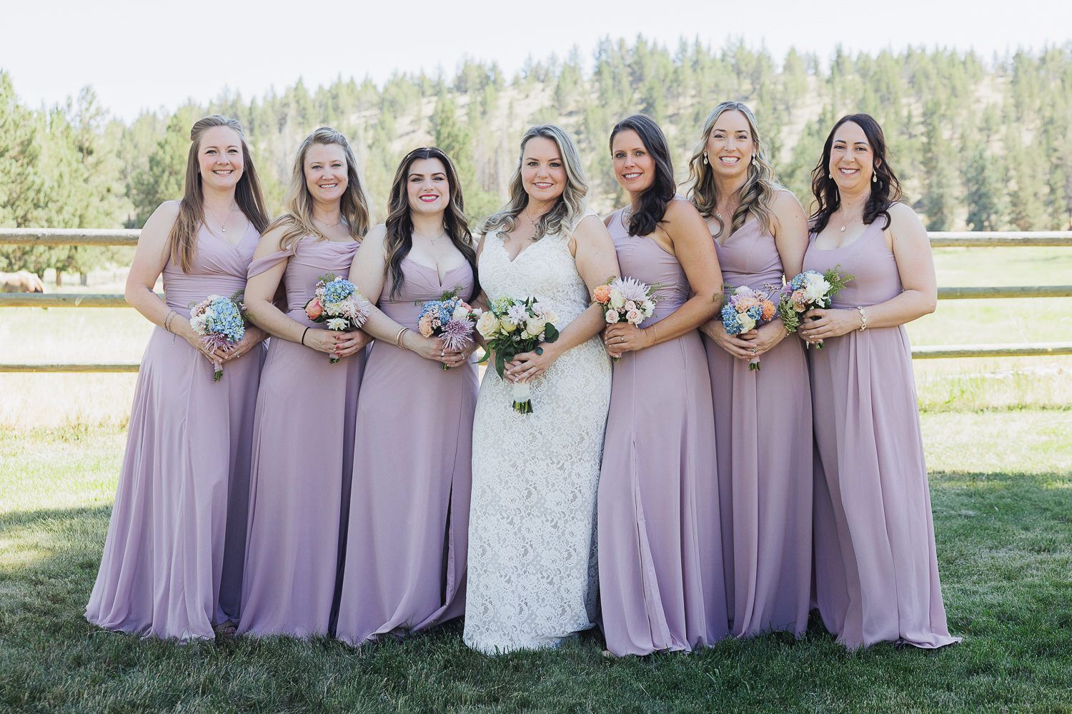 Wedding party in lavender bridesmaid dresses pose together outdoors against a scenic natural backdrop.
