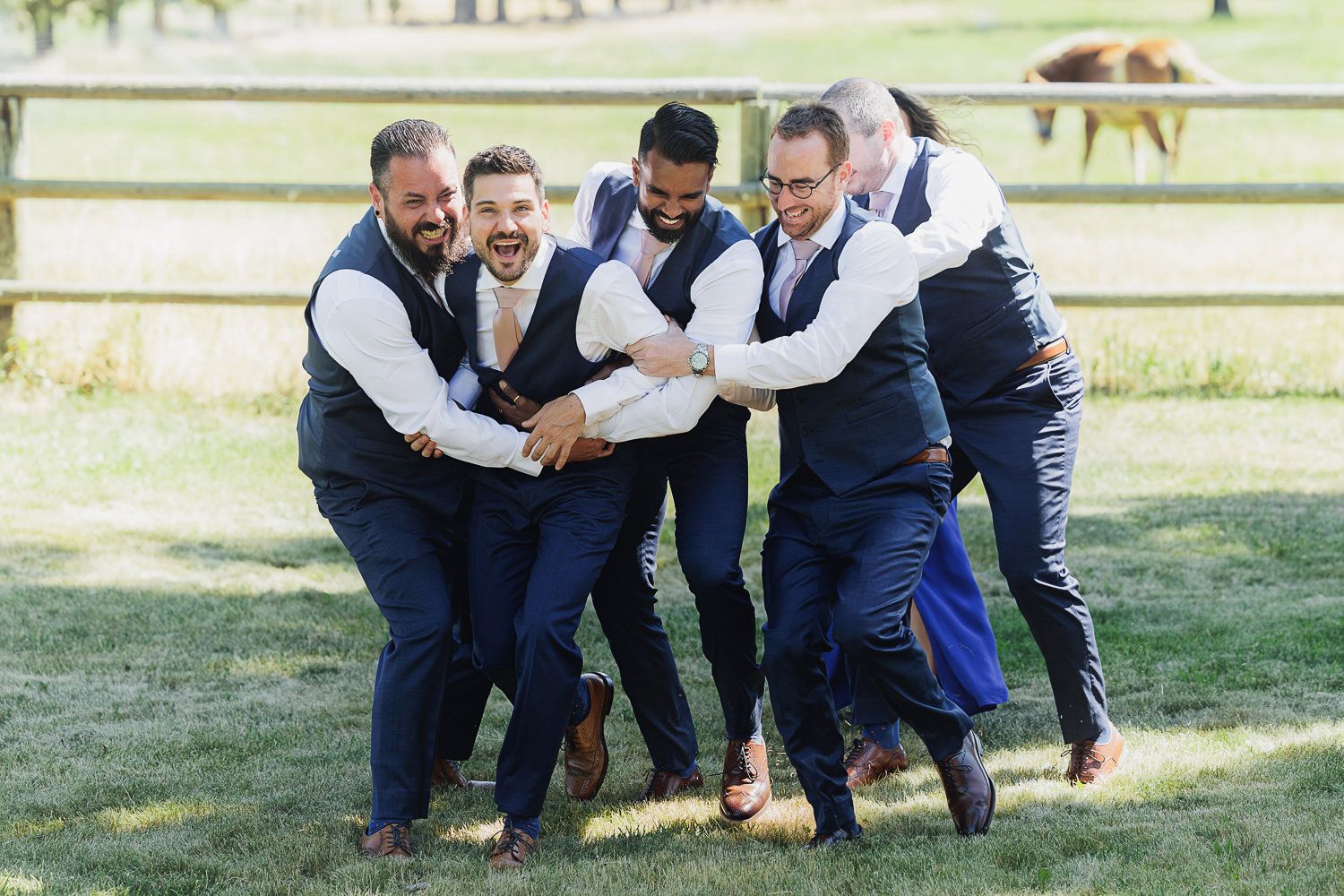 A group in matching navy vests and dress pants playfully huddle together on a grassy field with horses visible in background.