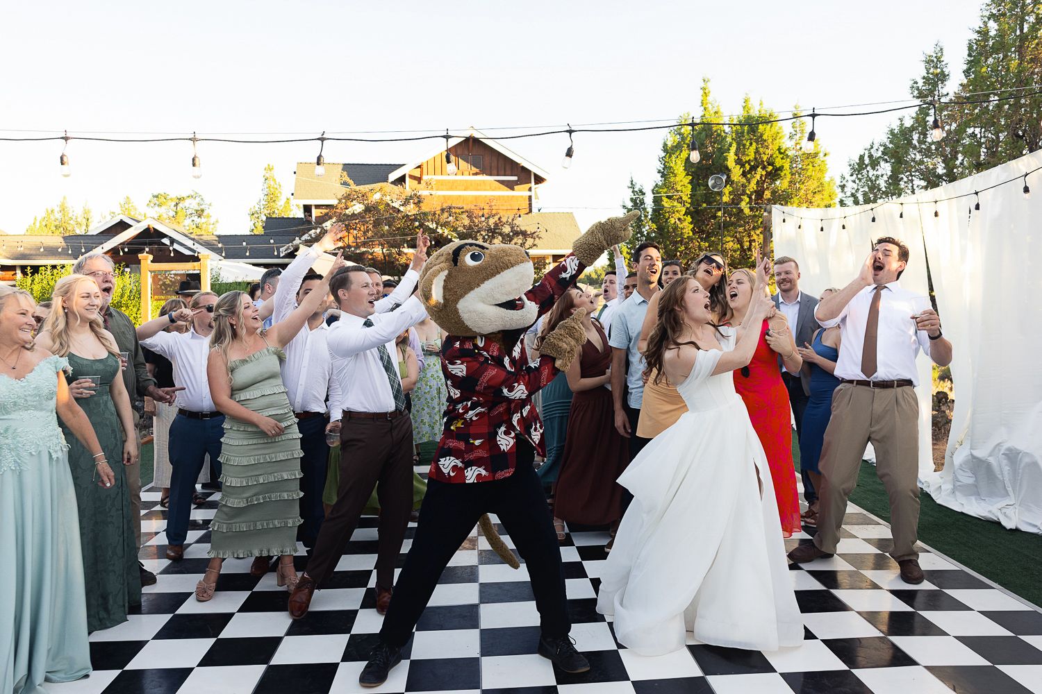 People dancing together on a black and white checkered dance floor at an outdoor wedding reception with string lights overhead.