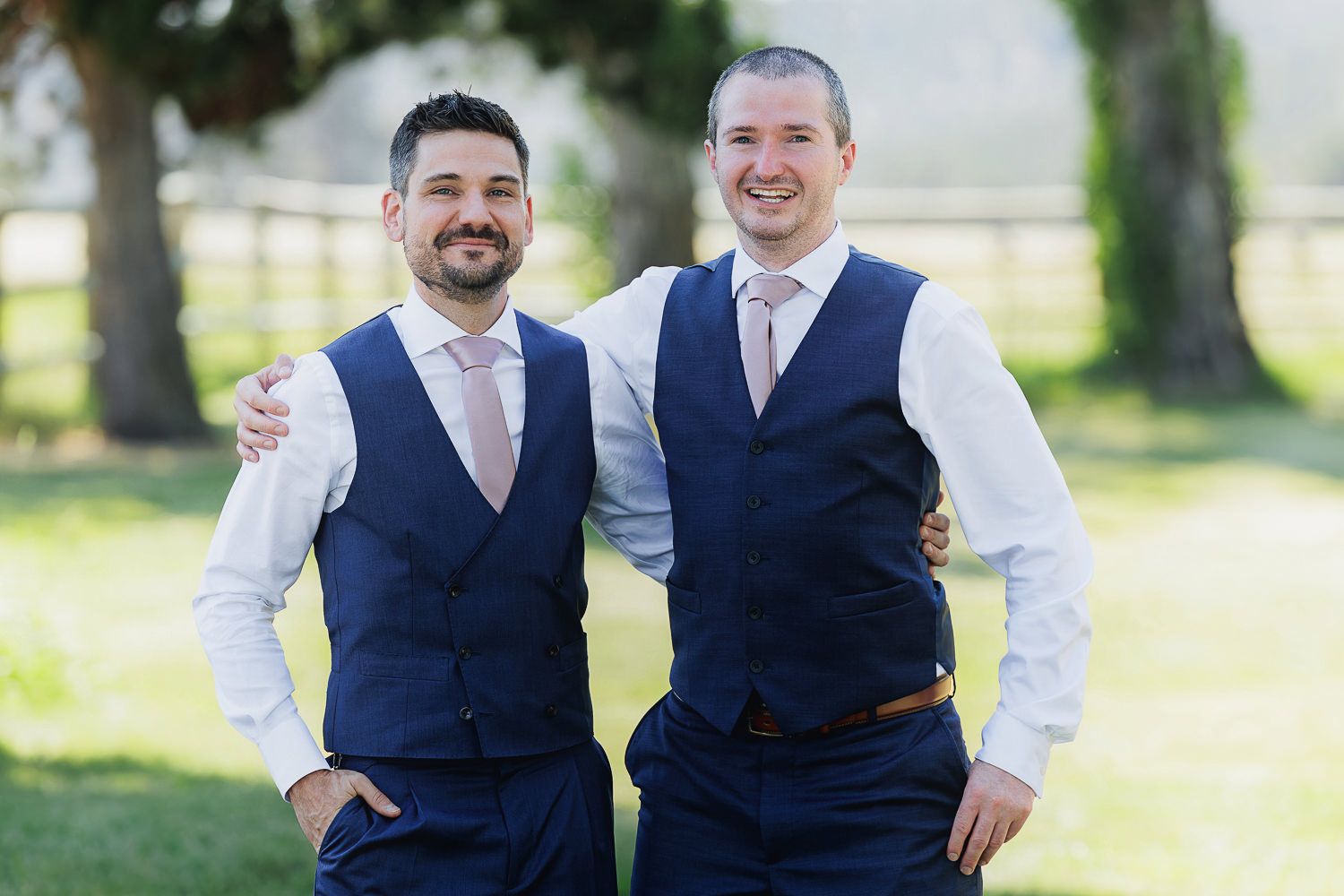 Two people wearing navy blue vests and light colored ties pose together on a grassy lawn.