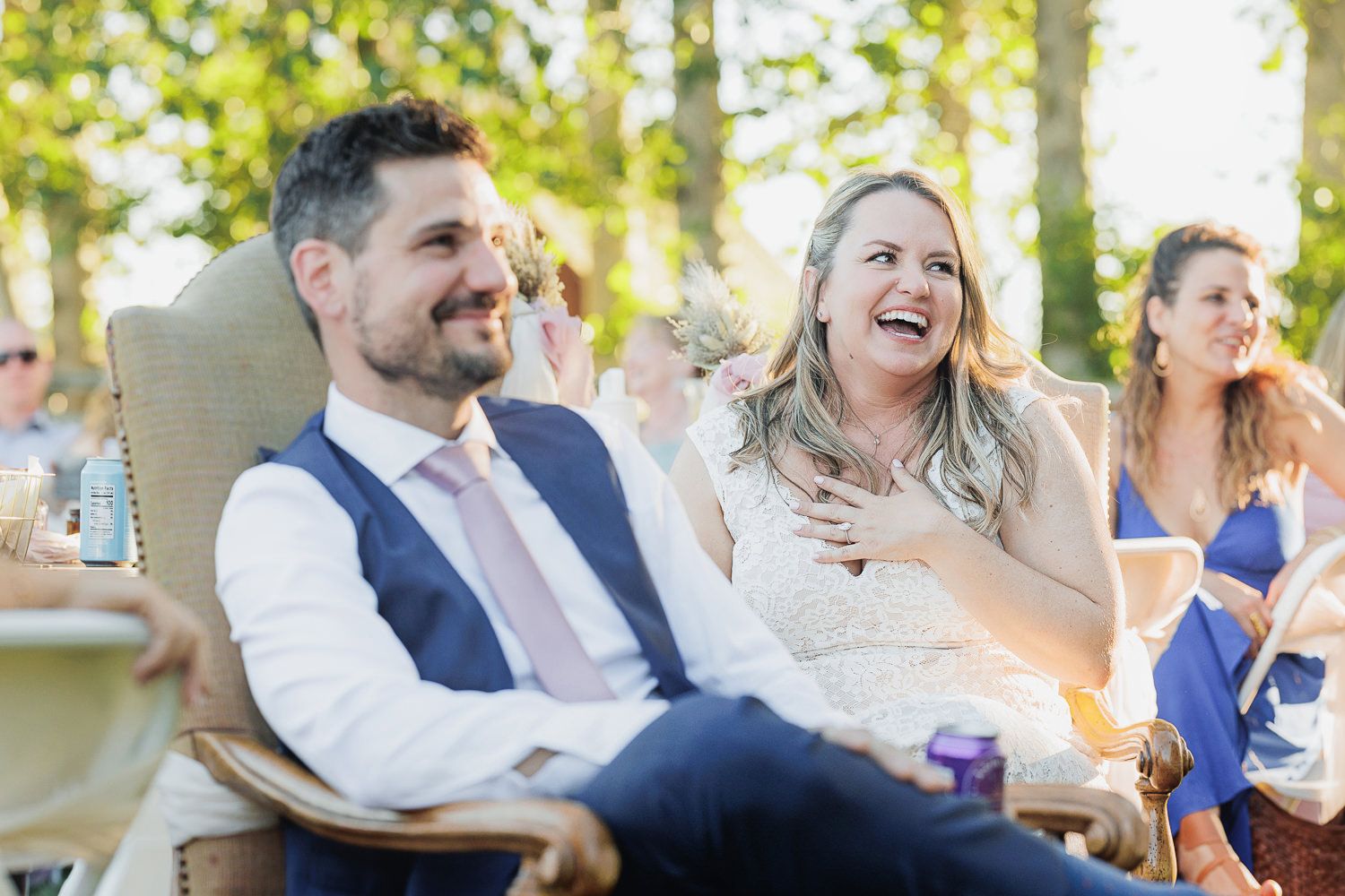 Wedding guests laugh together while sitting in outdoor chairs during a sunny reception celebration.