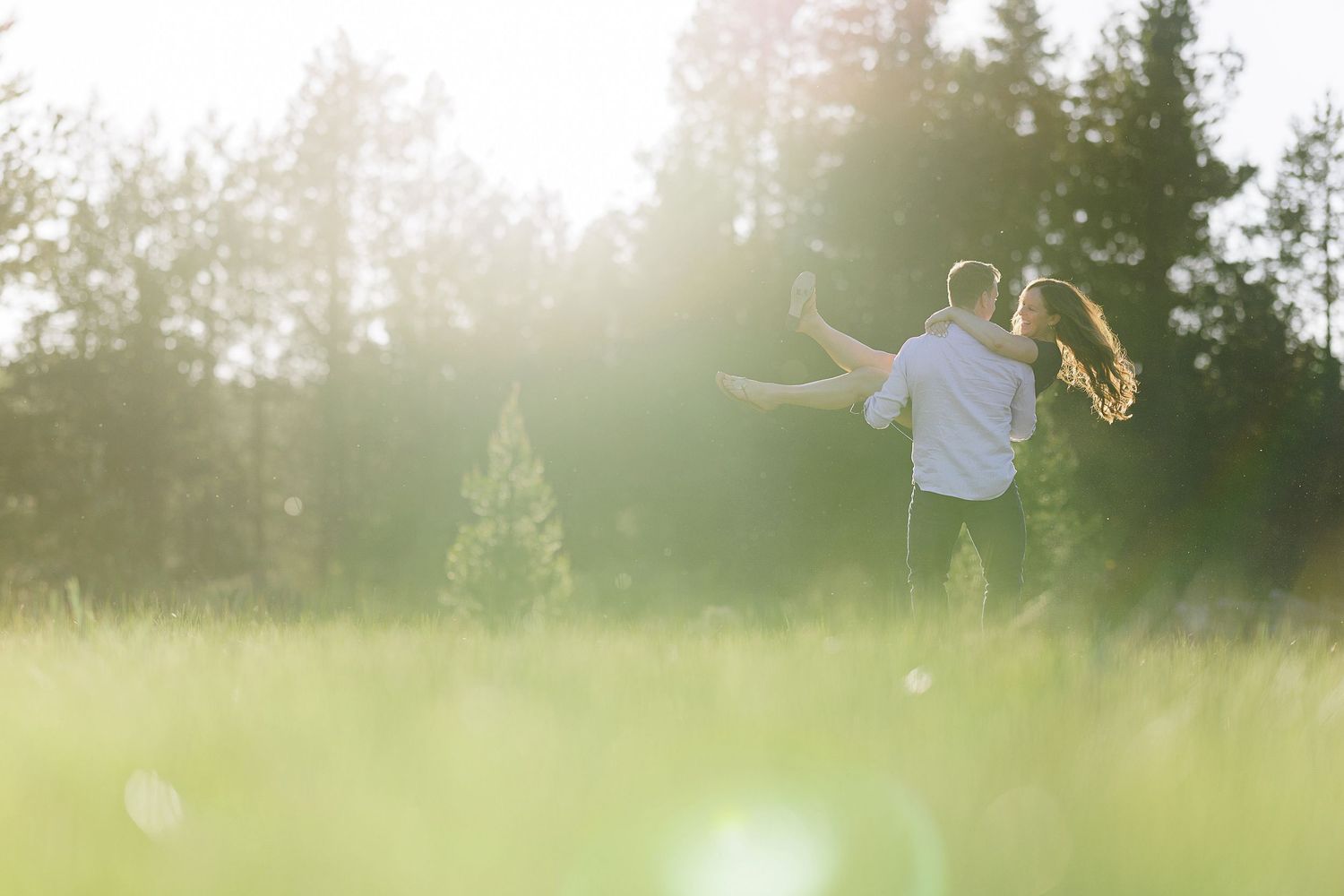 Two people running playfully through a sunlit meadow with evergreen trees in the background.