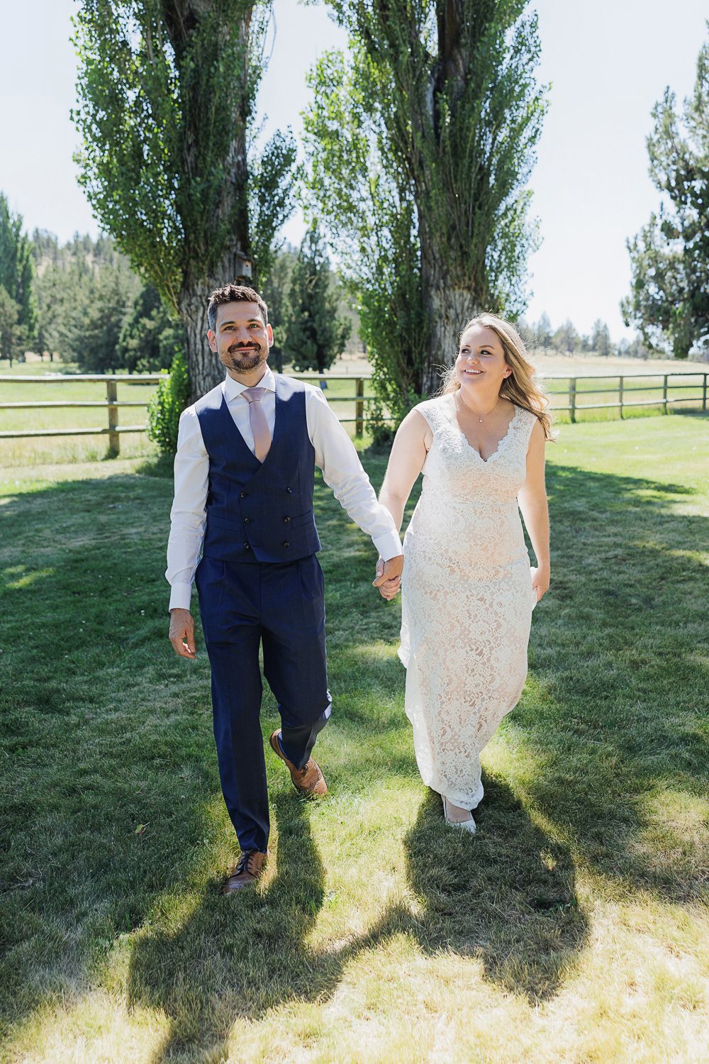 A couple walks hand in hand on a sunny lawn with tall poplar trees and wooden fencing in the background.