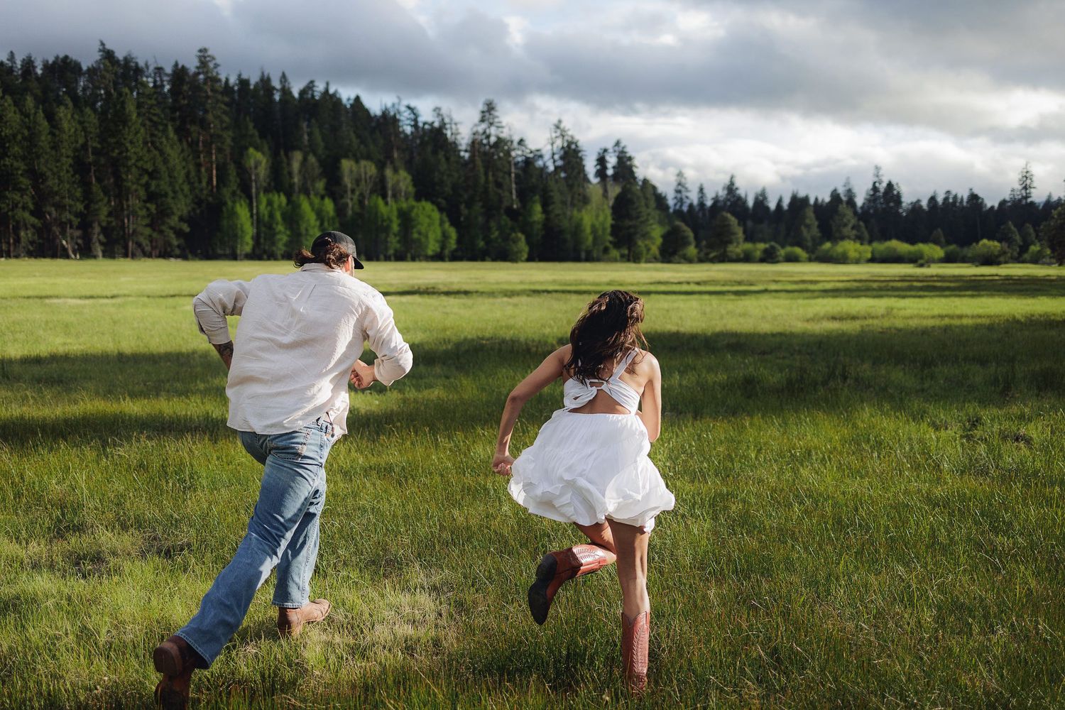 Two people run through a green meadow with boots on surrounded by evergreen forest under dramatic cloudy skies.