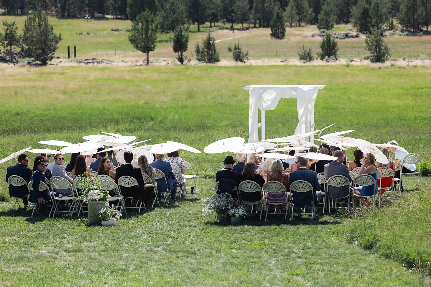 A panoramic view of an outdoor wedding setup with white chairs arranged on a green lawn surrounded by trees and mountains.