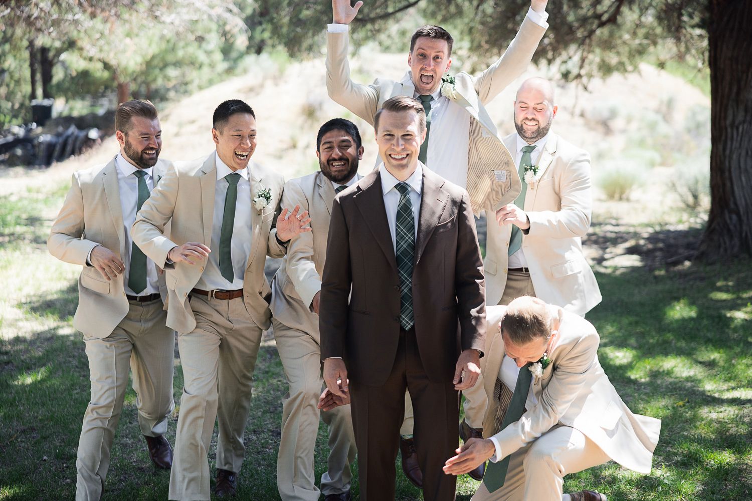 Wedding party in beige and brown suits joyfully celebrate together under trees in a garden setting.