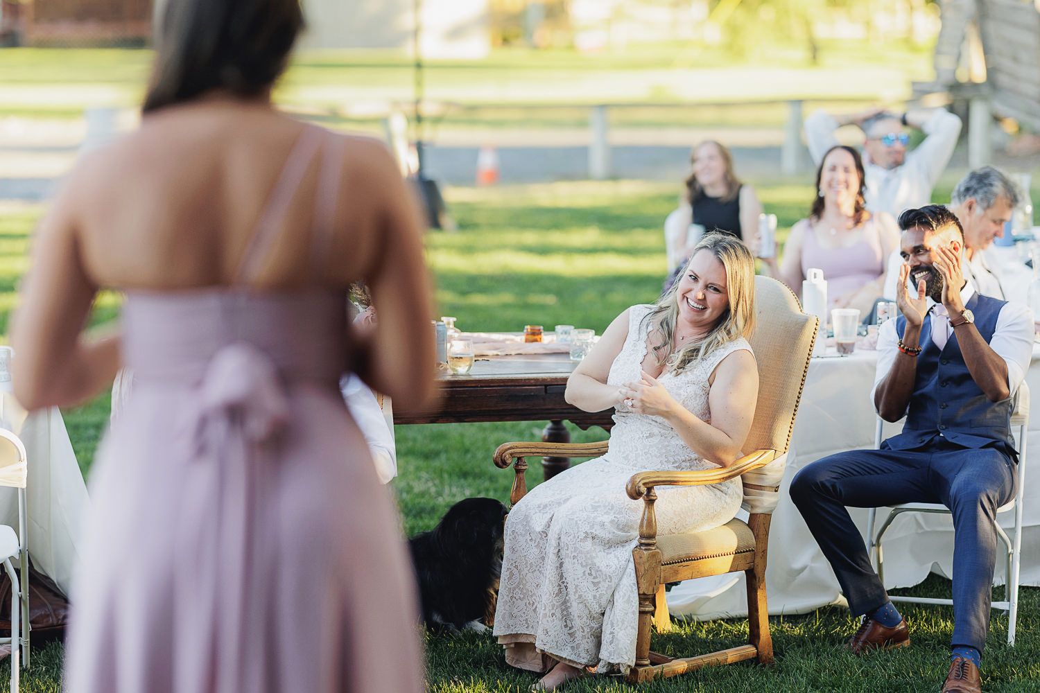 Wedding guests seated outdoors in a garden setting react emotionally as someone delivers a speech at a ceremony.