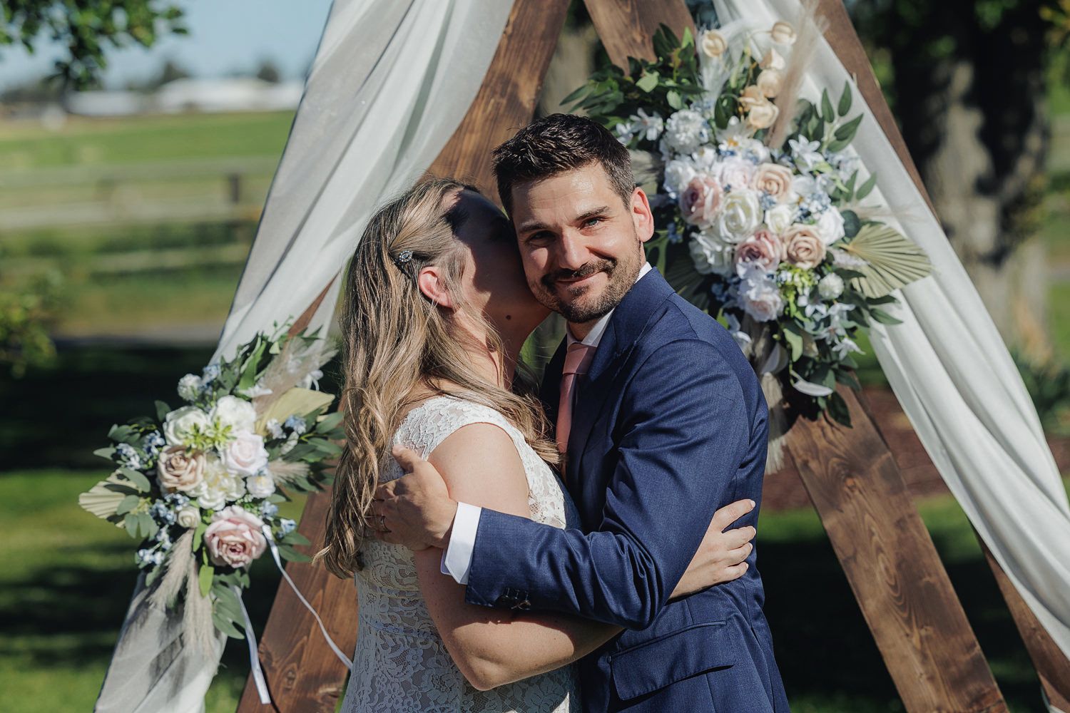 A joyful wedding couple embraces under a draped wooden arch decorated with pink and white floral arrangements outdoors.