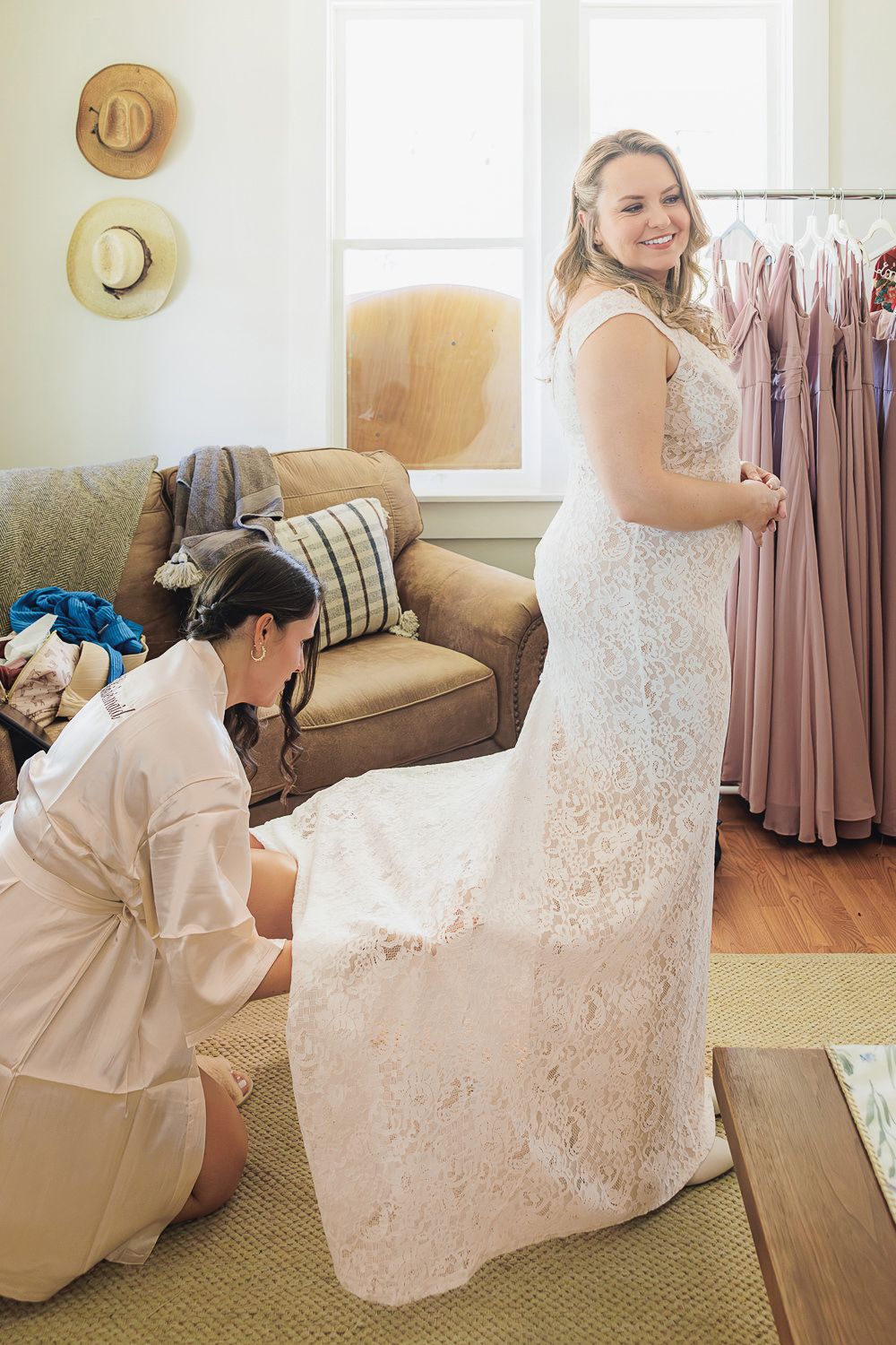 An attendant helps adjust a flowing white lace wedding dress while rosy bridesmaid dresses hang nearby.