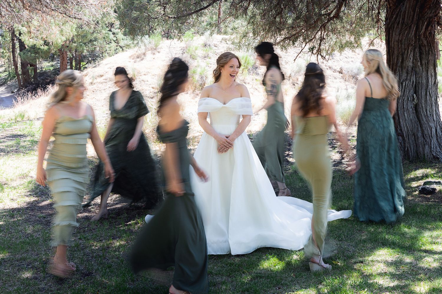 Wedding party members gather under shady trees for outdoor celebration photos.