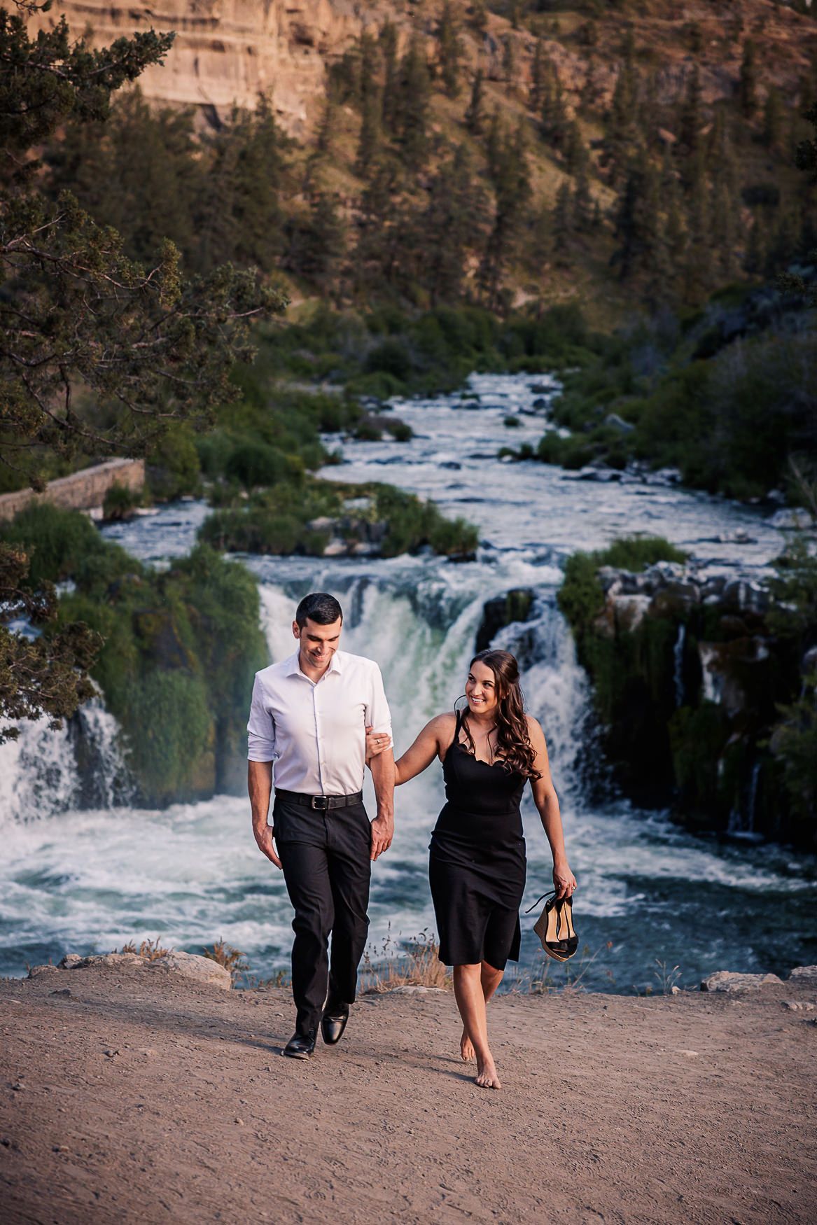 A couple walks hand in hand along a dirt path overlooking a dramatic waterfall and canyon landscape at sunset.