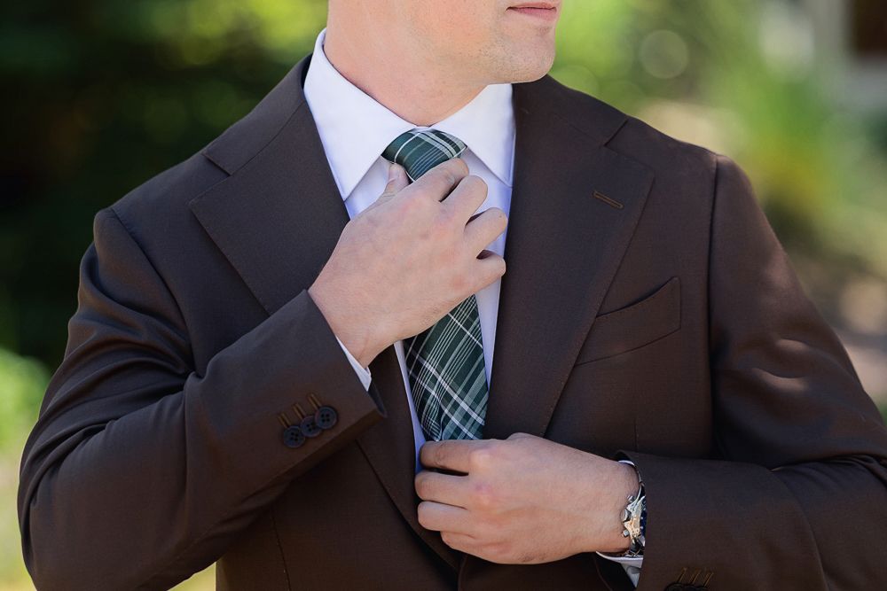 Professional in a brown business suit adjusts striped necktie against blurred green outdoor background.