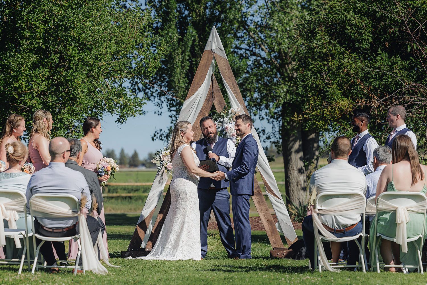 Outdoor wedding ceremony in front of a rustic wooden triangle arch with guests seated on white chairs on a sunny day.