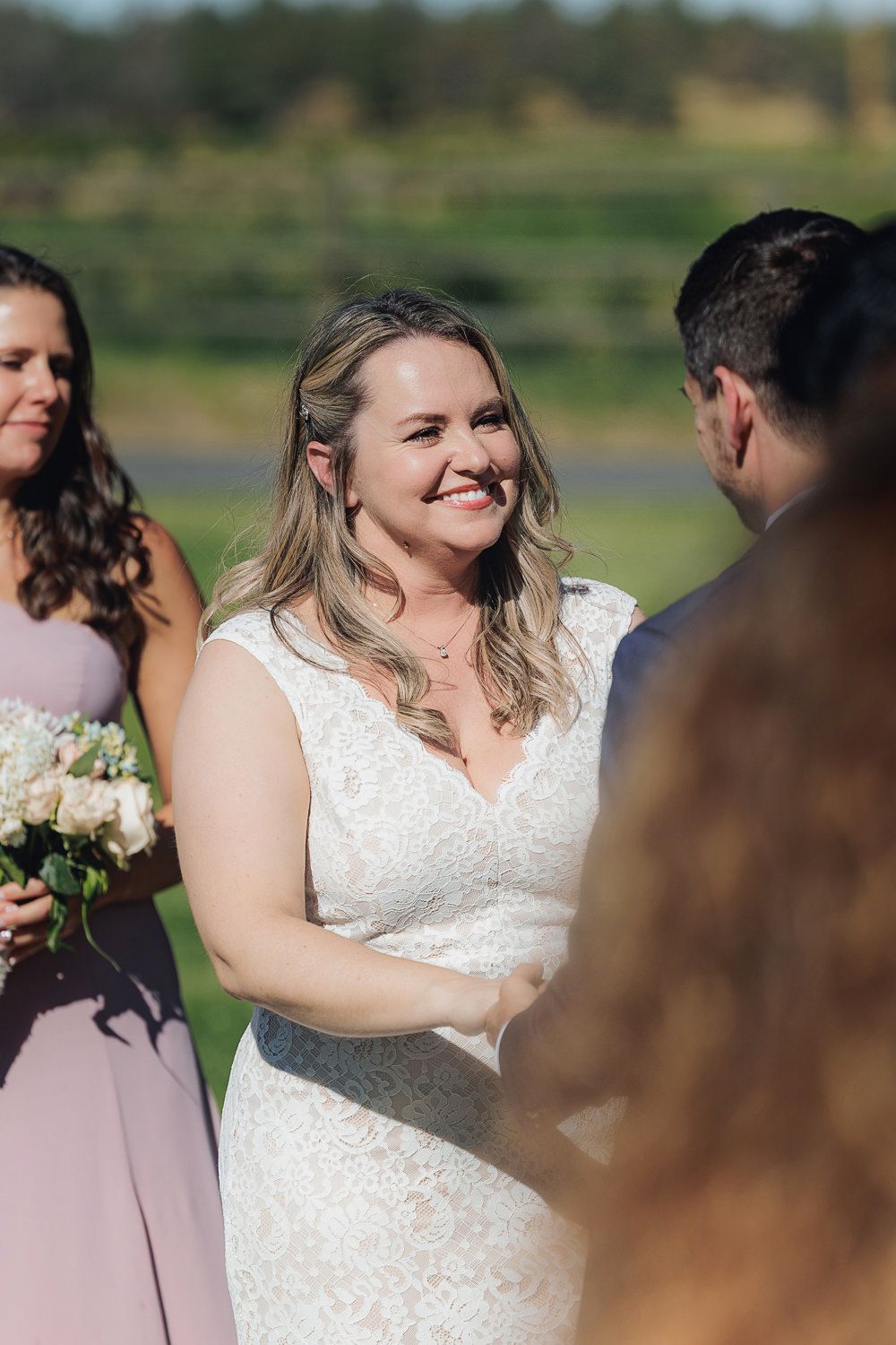 A wedding ceremony with a bride wearing a beautiful lace dress standing with attendants in mauve gowns outdoors.