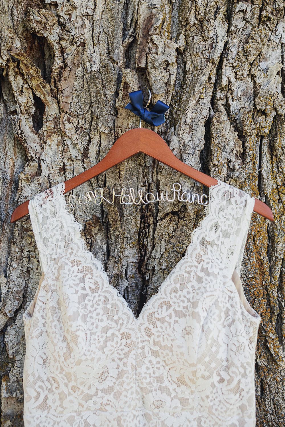 A white lace wedding dress hangs on a wooden hanger against a tree bark background with a blue detail at the top.