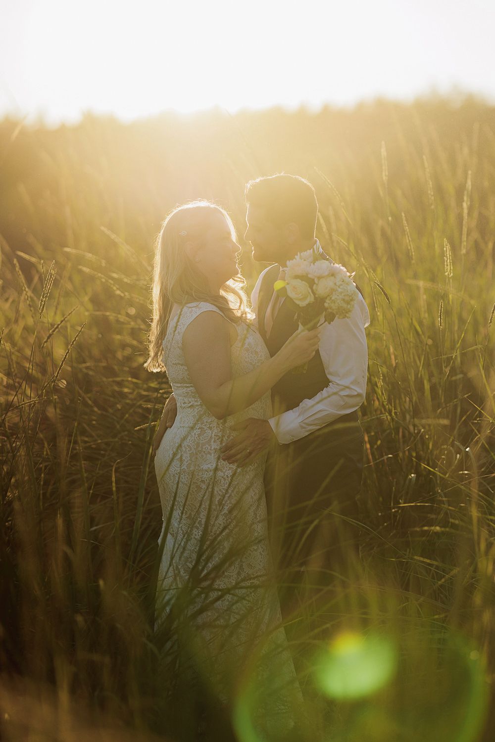 A romantic couple in white wedding attire embraces in a golden sunlit meadow of tall grass during sunset.
