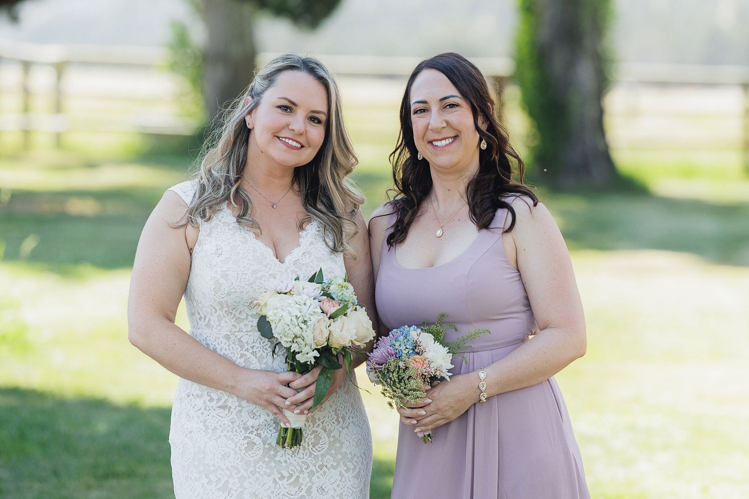 A bride in a lace wedding dress and bridesmaid in mauve holding white flower bouquets pose outdoors on a sunny day.