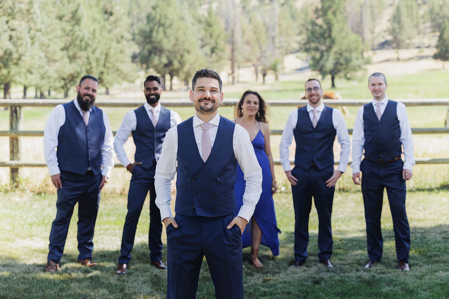 Wedding party wearing coordinated navy blue vests poses together outdoors in front of a rustic wooden fence.