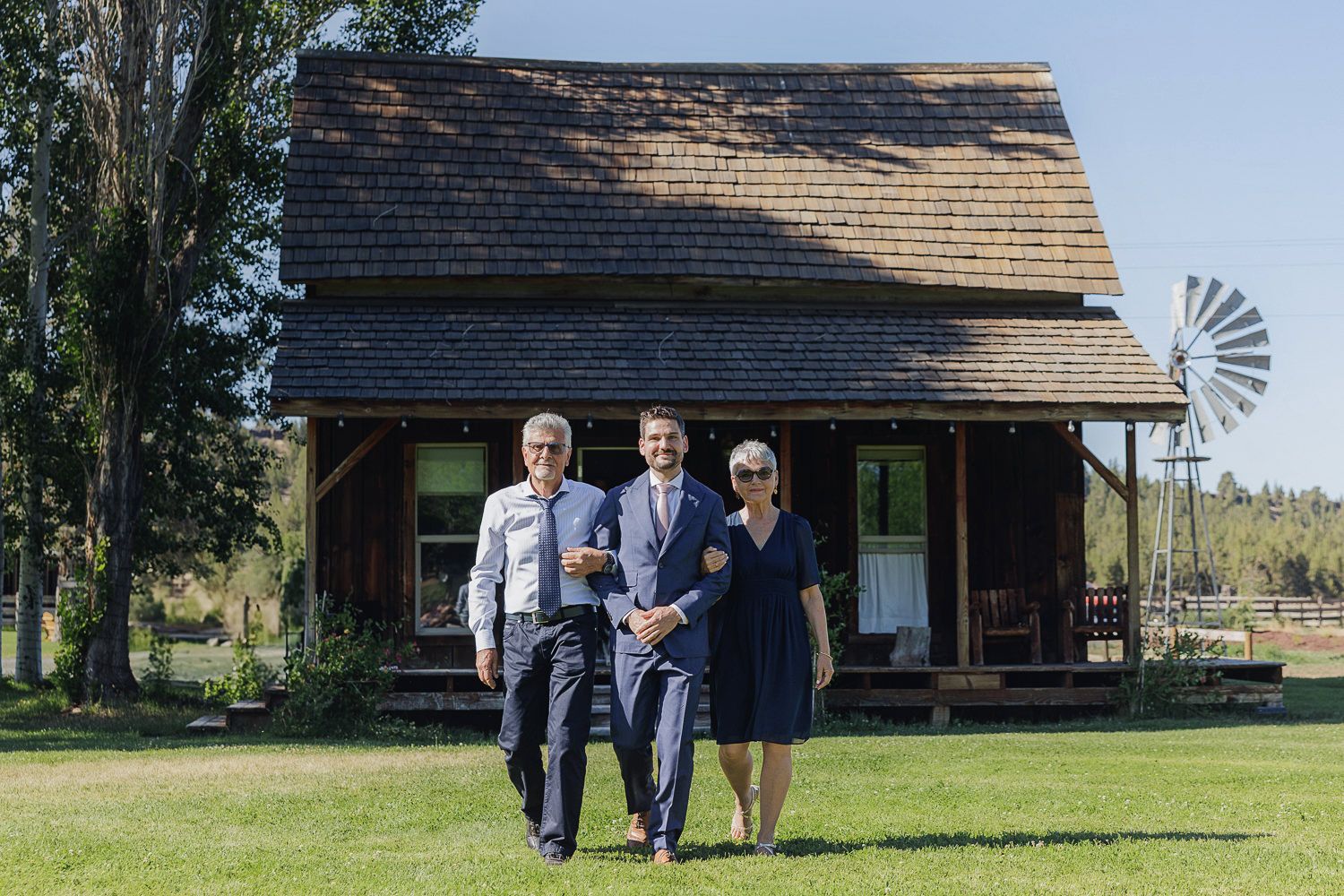 A group poses in front of a rustic log cabin with a vintage windmill visible in the background.