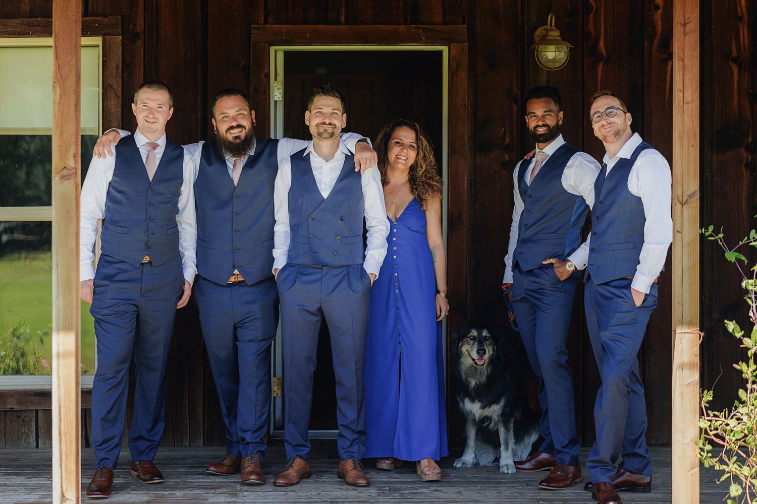 Wedding party in matching navy blue vests and pants poses with a black dog on a wooden porch.