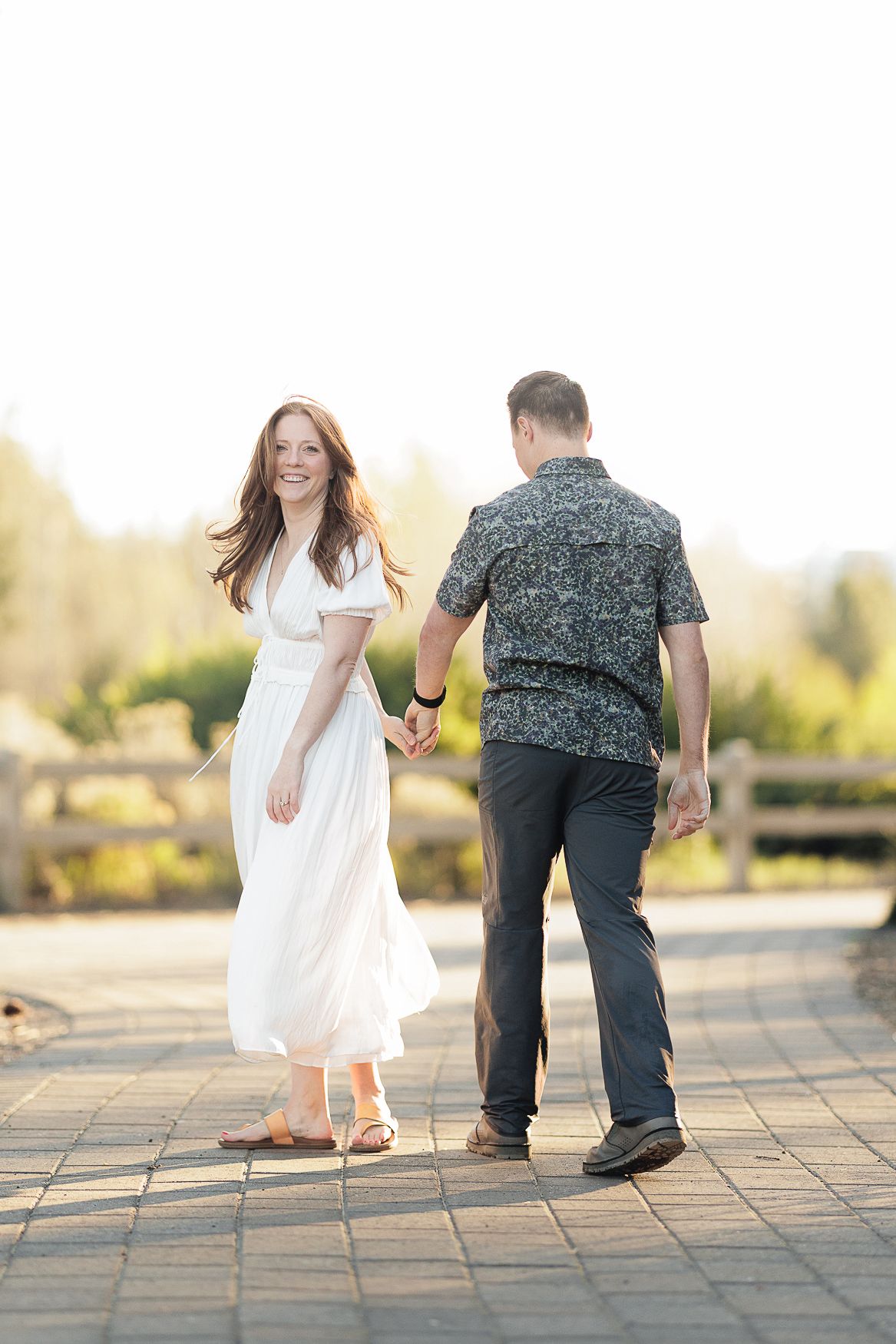 Joyful couple walks hand-in-hand on stone patio with soft golden sunset lighting in background.