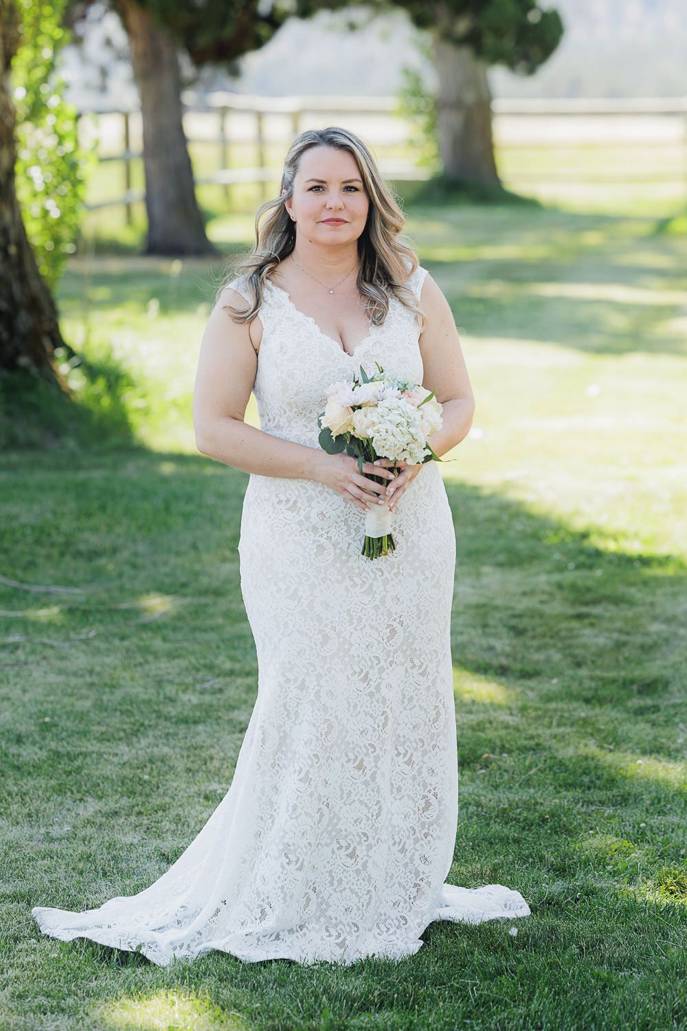 A bride in a fitted lace wedding gown holds a white bouquet while standing in a sun-dappled garden setting.