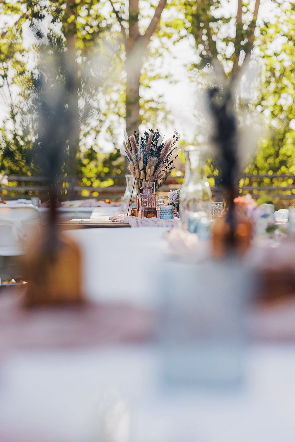 Rustic outdoor table setting with centerpiece bouquet seen through blurred foreground elements in warm sunlit garden.