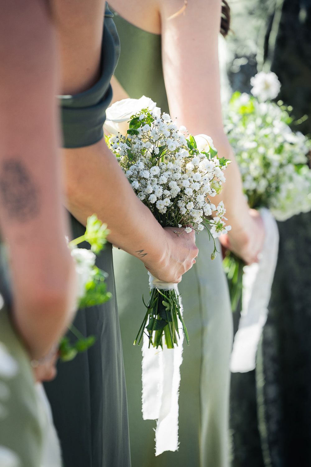A bridesmaid in a sage green dress holds a delicate bouquet of white baby's breath flowers tied with a white ribbon.