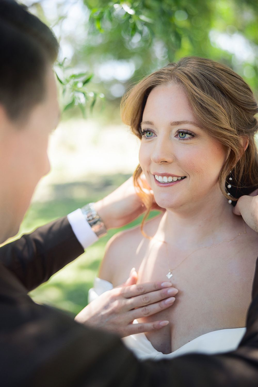 A bride in a white wedding dress smiles while someone fixes her hair at an outdoor ceremony.