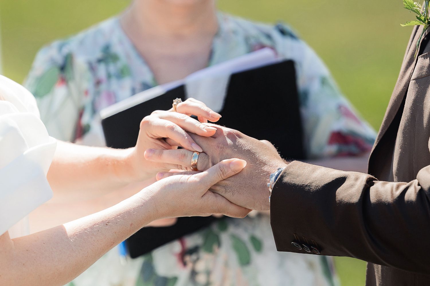 Close up view of hands exchanging rings during outdoor wedding ceremony with floral dress in background.