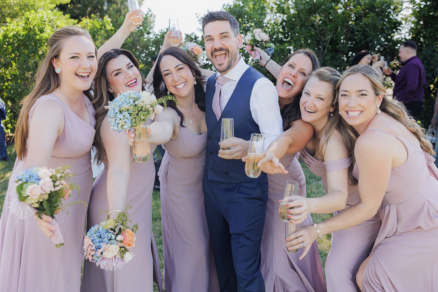 A cheerful wedding party in pink bridesmaid dresses poses together outdoors while holding colorful bouquets.