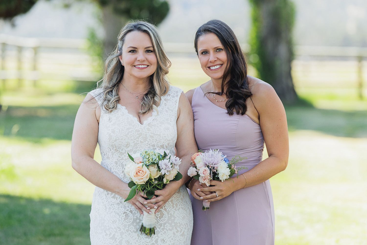 Two women in wedding attire pose with bouquets in a sunny outdoor garden setting.