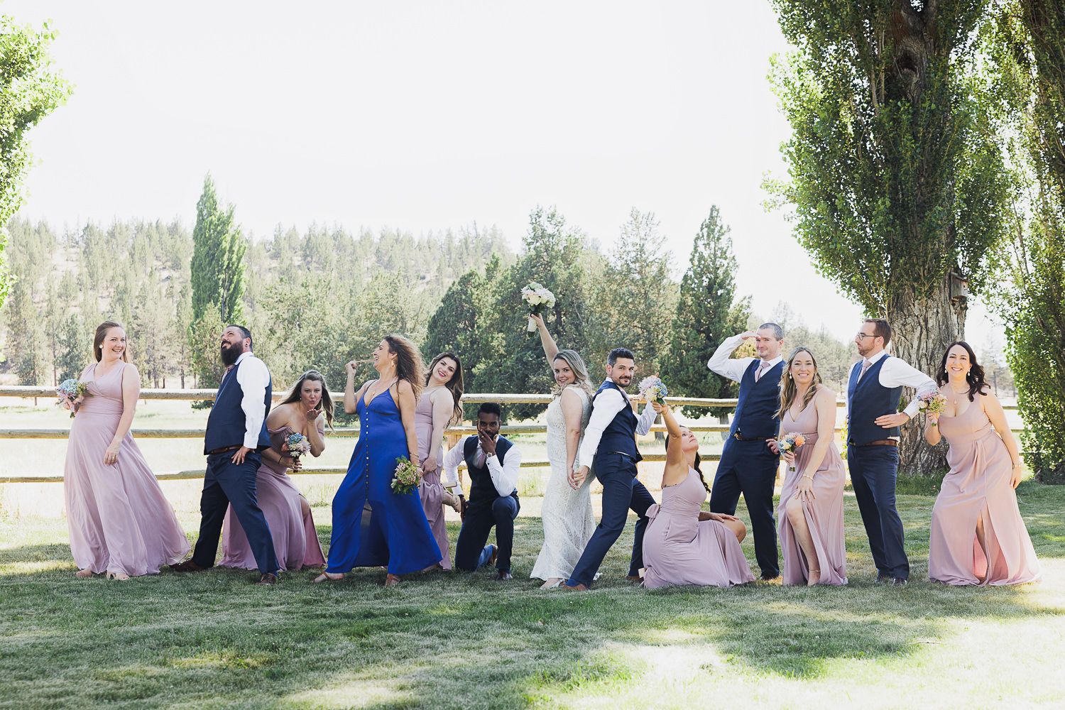 Wedding party in colorful attire poses playfully in a row for outdoor photo session against scenic natural backdrop.