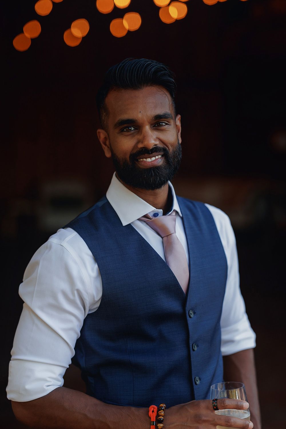 Well-dressed person in navy blue vest and white dress shirt smiling while holding drink glass against dark background.