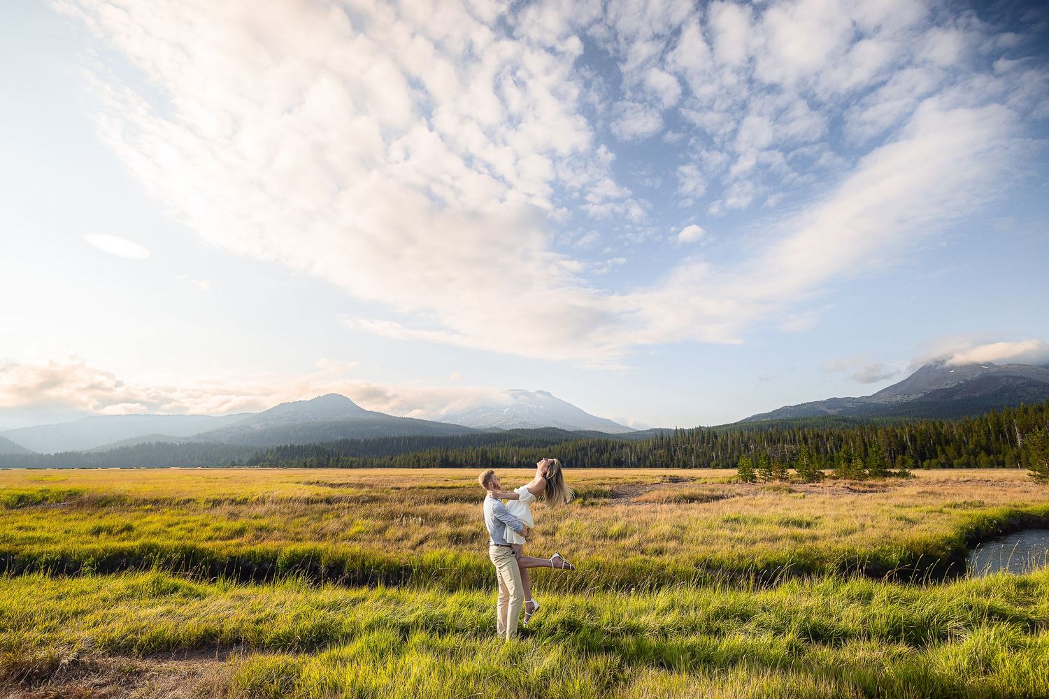 A couple stands in a golden meadow with mountains and cloudy skies in the backdrop during golden hour.