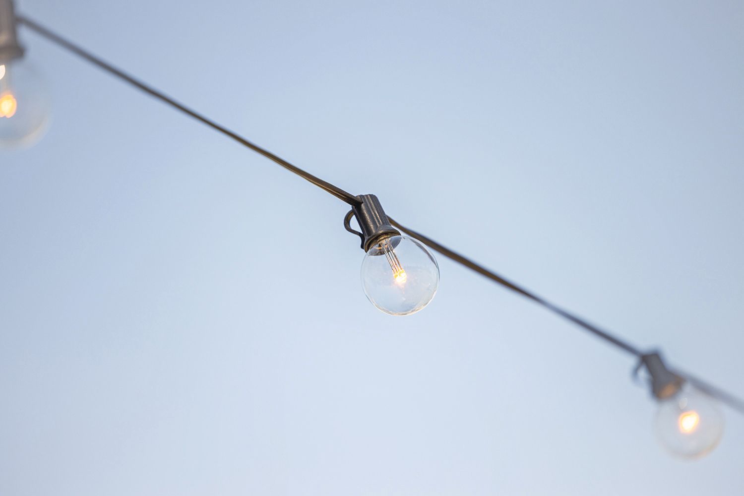 String of outdoor bistro lights with round bulbs against a pale blue sky.