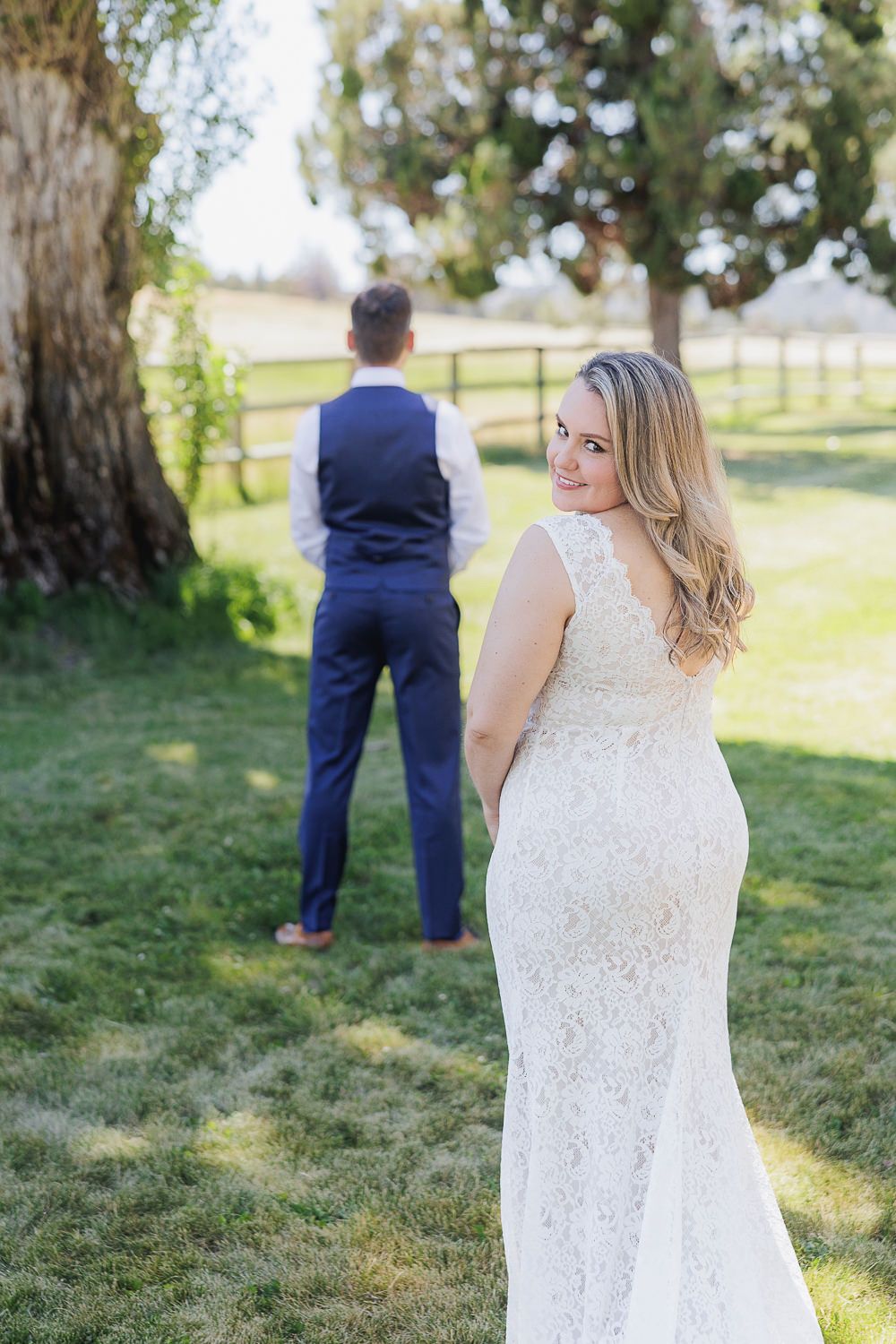 A bride in a white lace dress stands on grass while looking back over her shoulder at a distance.