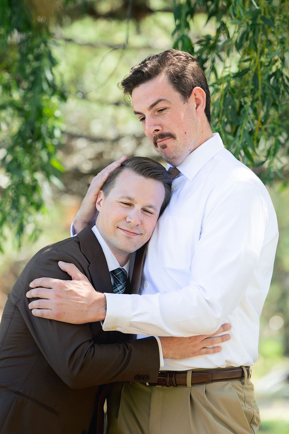 Two people share a tender embrace under willow tree branches in a garden setting.