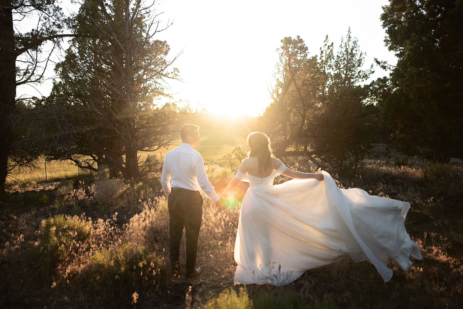A couple holding hands walks through a golden sunset in a natural outdoor setting as a wedding dress train flows behind them.