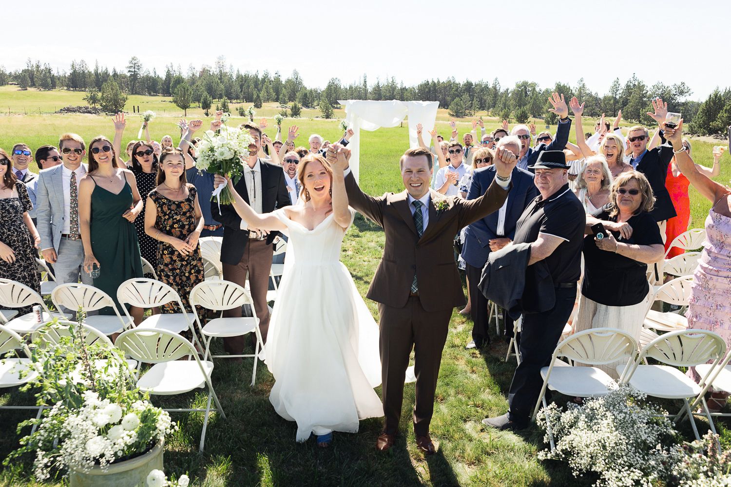 Wedding guests seated in white chairs during an outdoor ceremony with mountains and trees in the background.